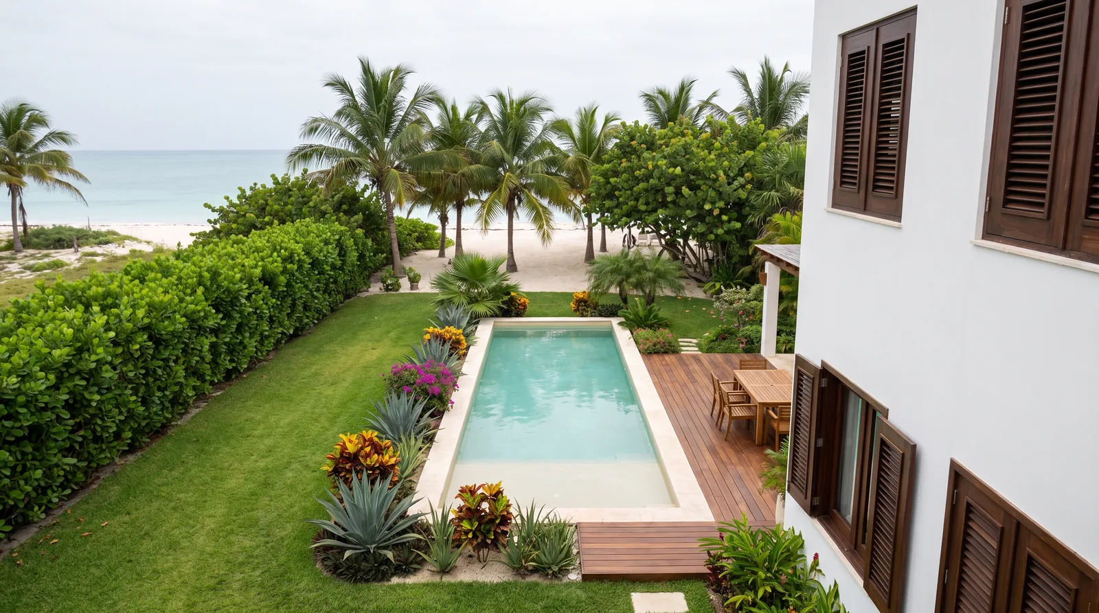 Jardín detrás de una casa blanca en la costa de Yucatán: alberca, deck de madera, riego encendido, palmeras y al fondo la playa y el mar