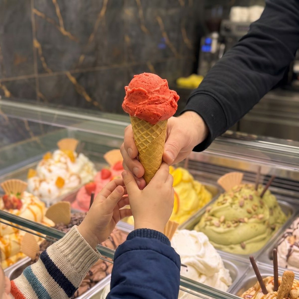 Eine Hand reicht eine Eiswaffel über die Theke an zwei Kinderhände