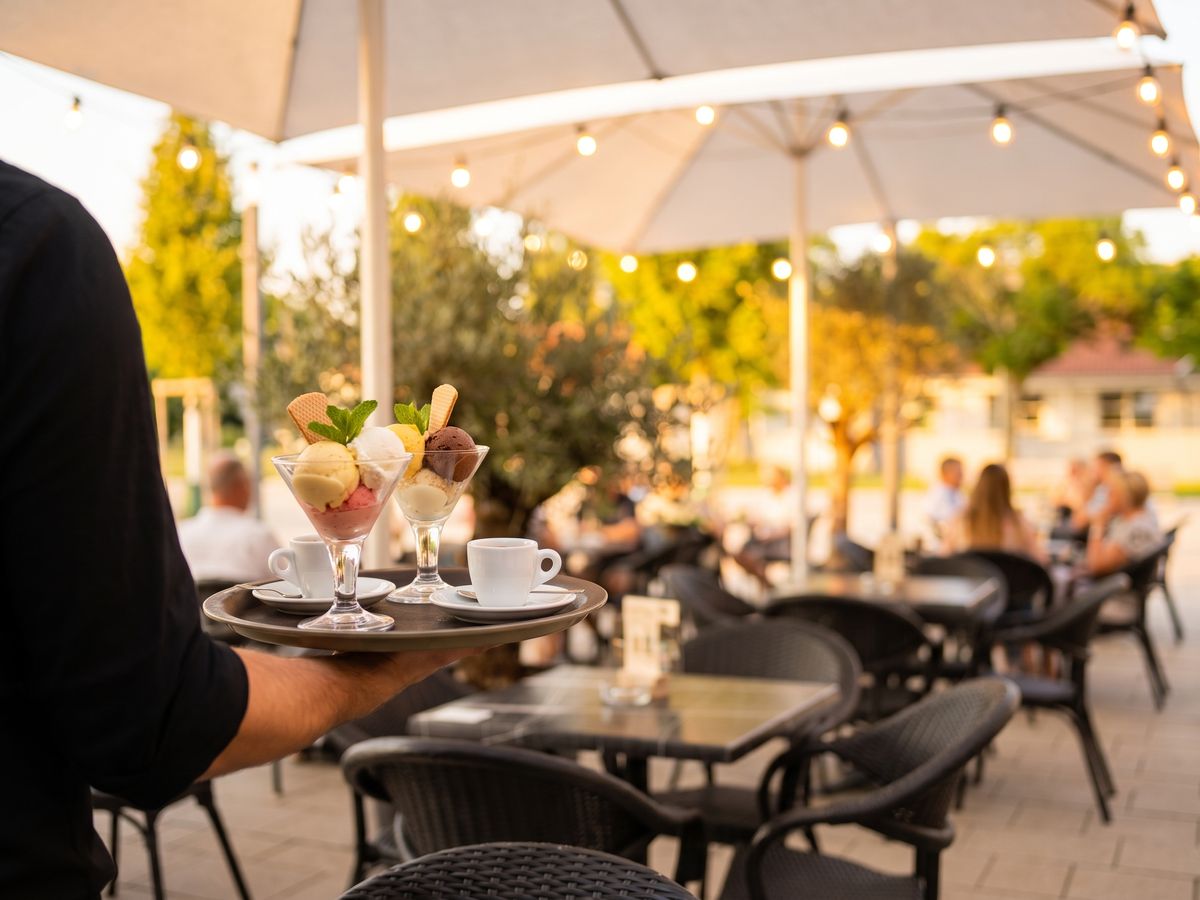Kellner trägt ein Tablett mit Eisbechern und Espresso über die Terrasse mit Lichterketten