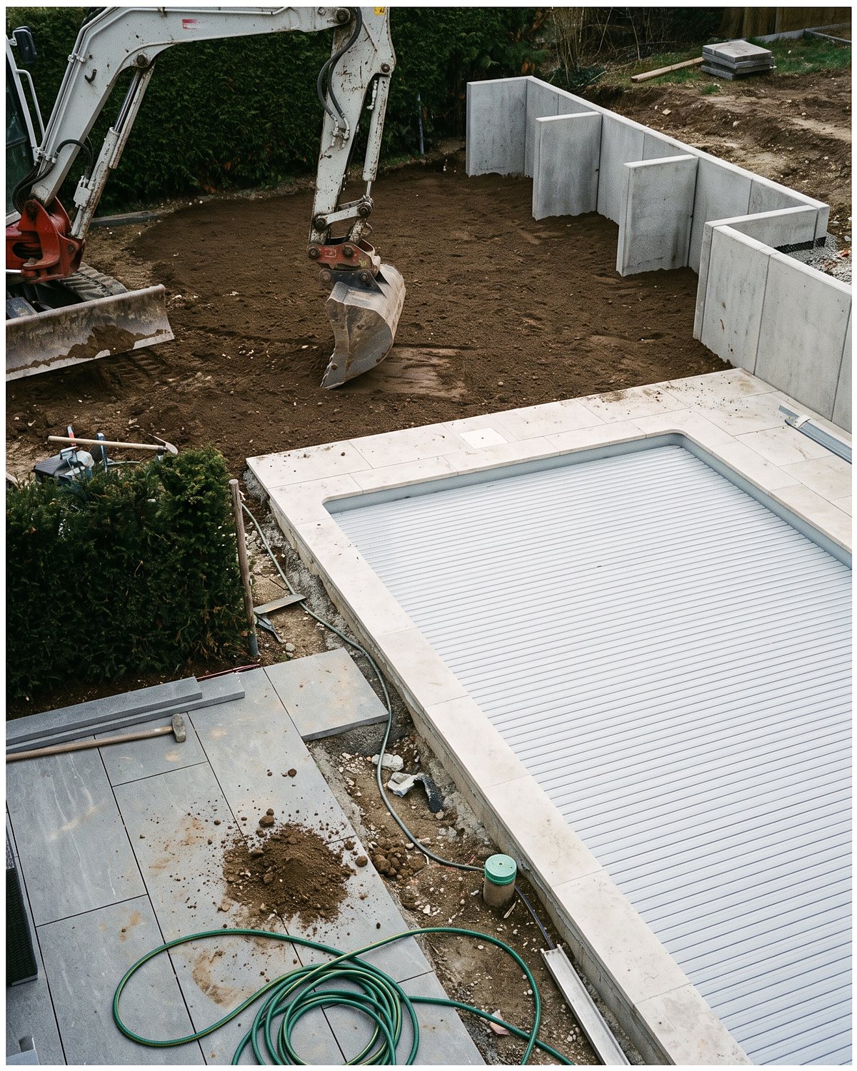 Fertige Poolanlage mit Abdeckung, Terrassenplatten und Randeinfassung, im Hintergrund arbeitet der Bagger am Erdreich