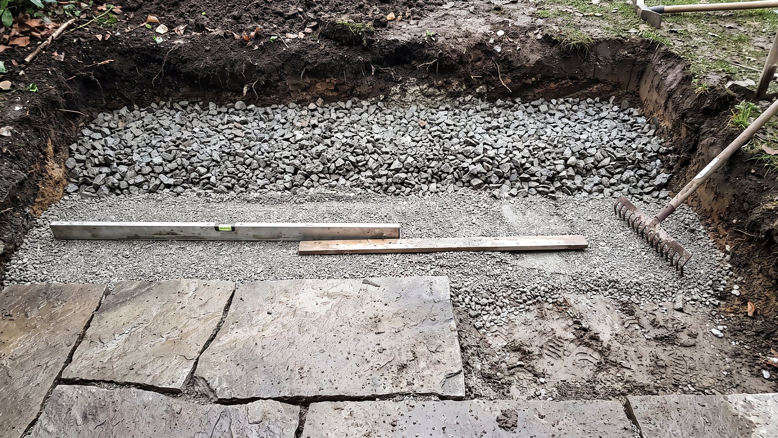 Unterbau einer Terrasse von Gartenbau Mustermann: Schotter, Splittbett und die ersten verlegten Platten, daneben Wasserwaage und Rechen